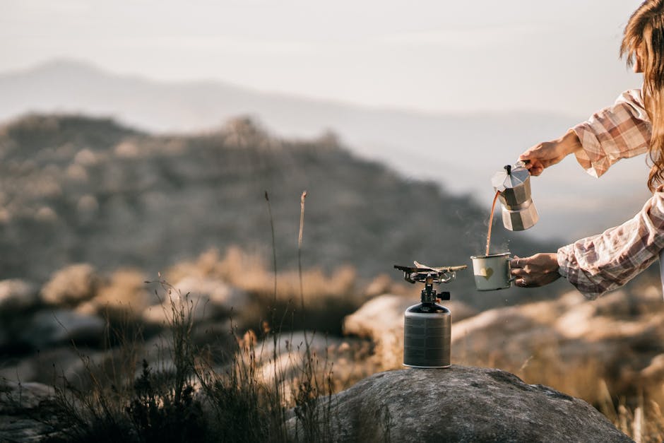 A person brews coffee using a portable stove in a scenic mountain setting during sunrise.
